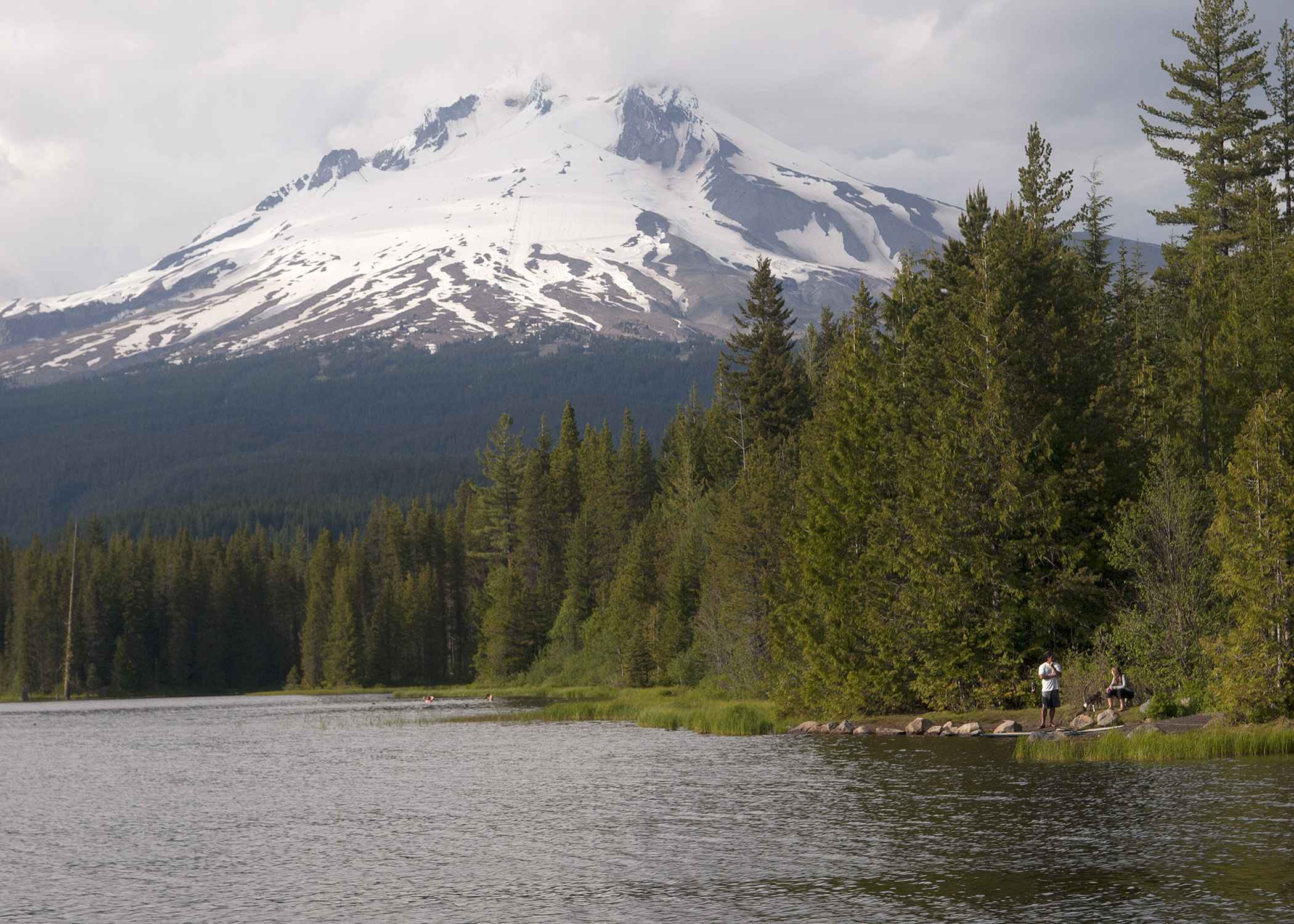 Mount Hood rising above Trillium Lake, with forest and two visitors near the shoreline.